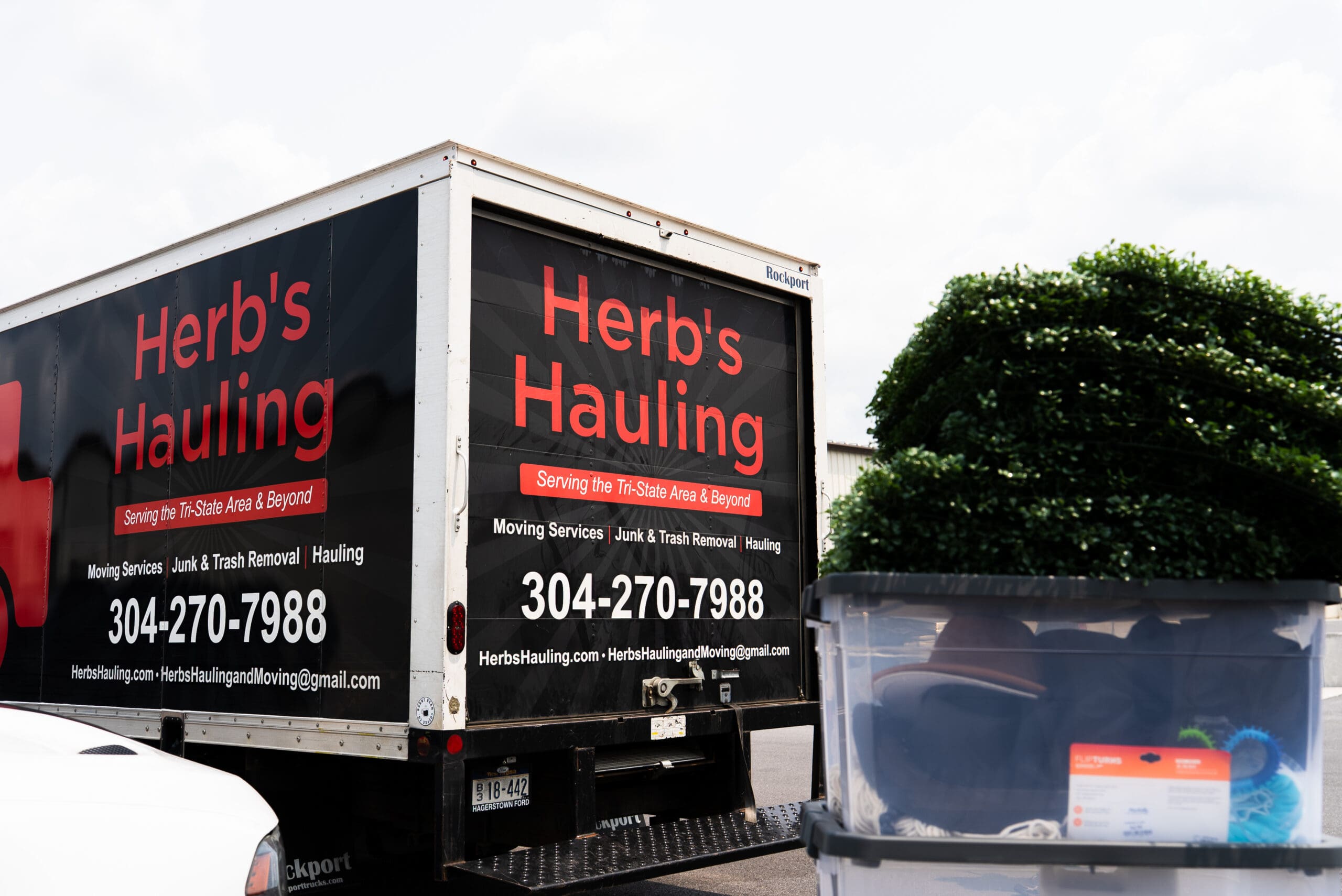 A black commercial hauling truck with red Herb's Hauling signage parked on the side of a road with trees and another vehicle visible.