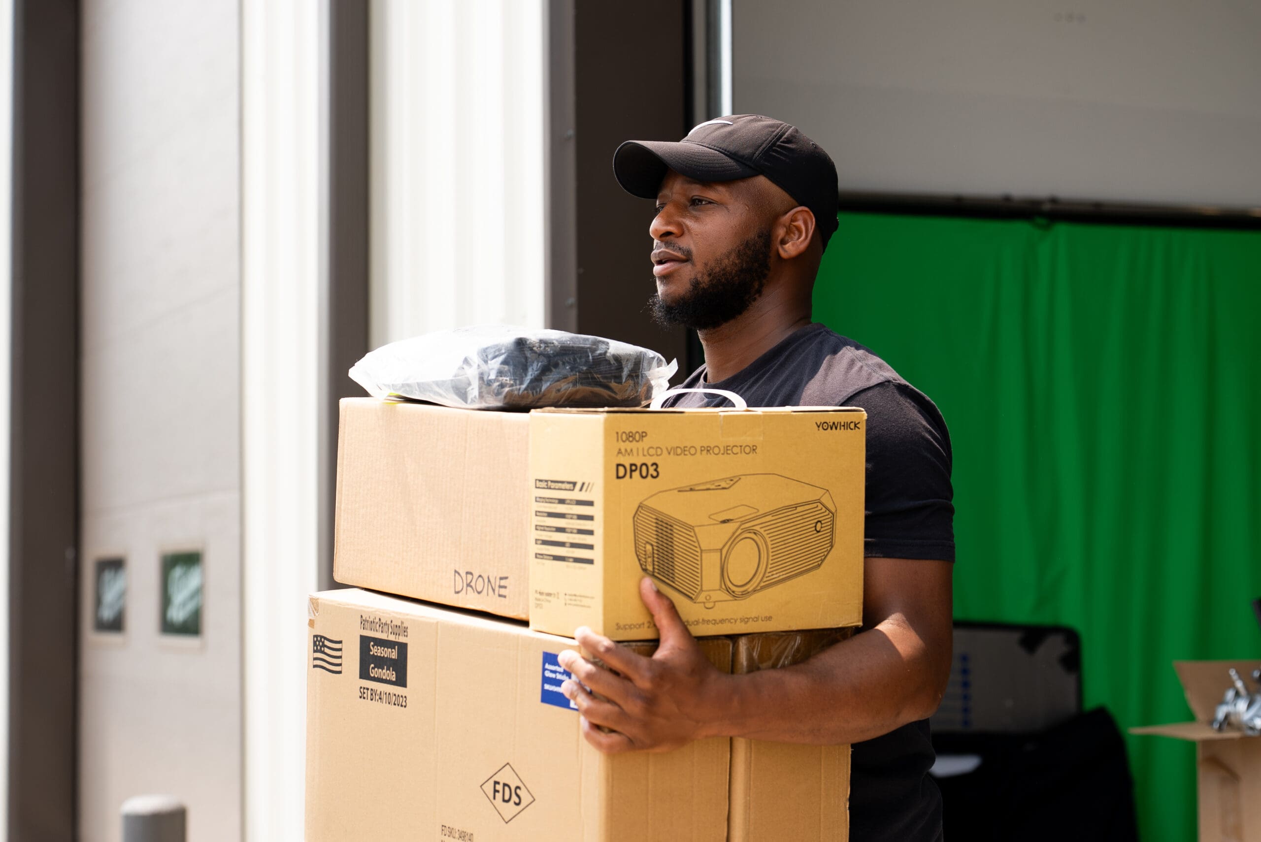A man wearing a cap holds a large cardboard box in an indoor workshop or storage space with green walls and white columns.