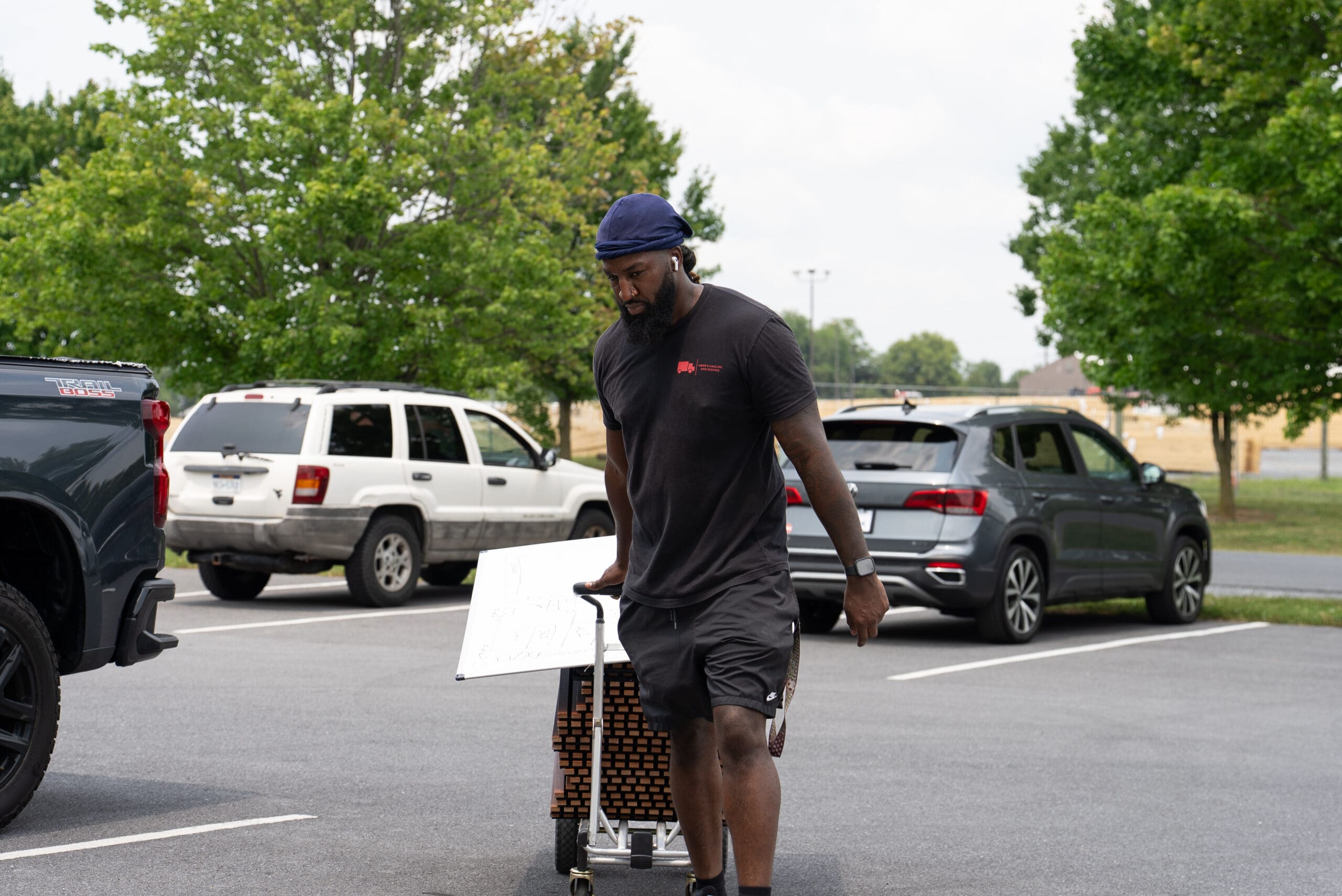 A person in dark clothing rides a bicycle through a suburban parking lot lined with parked vehicles and trees on a daytime street.