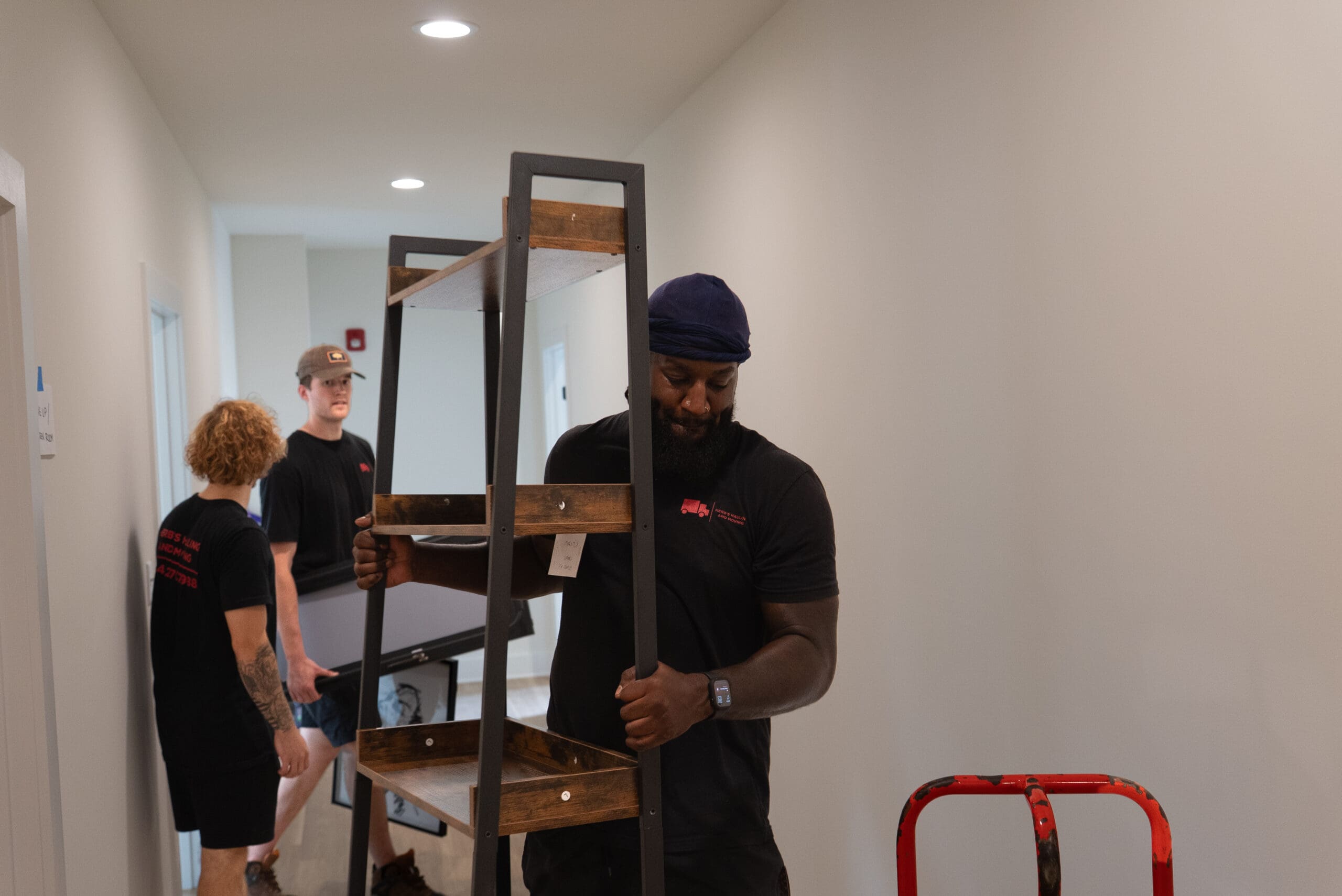 A workshop instructor in black clothing demonstrates a furniture assembly or installation task to a small group of participants in a bright, clean interior space.