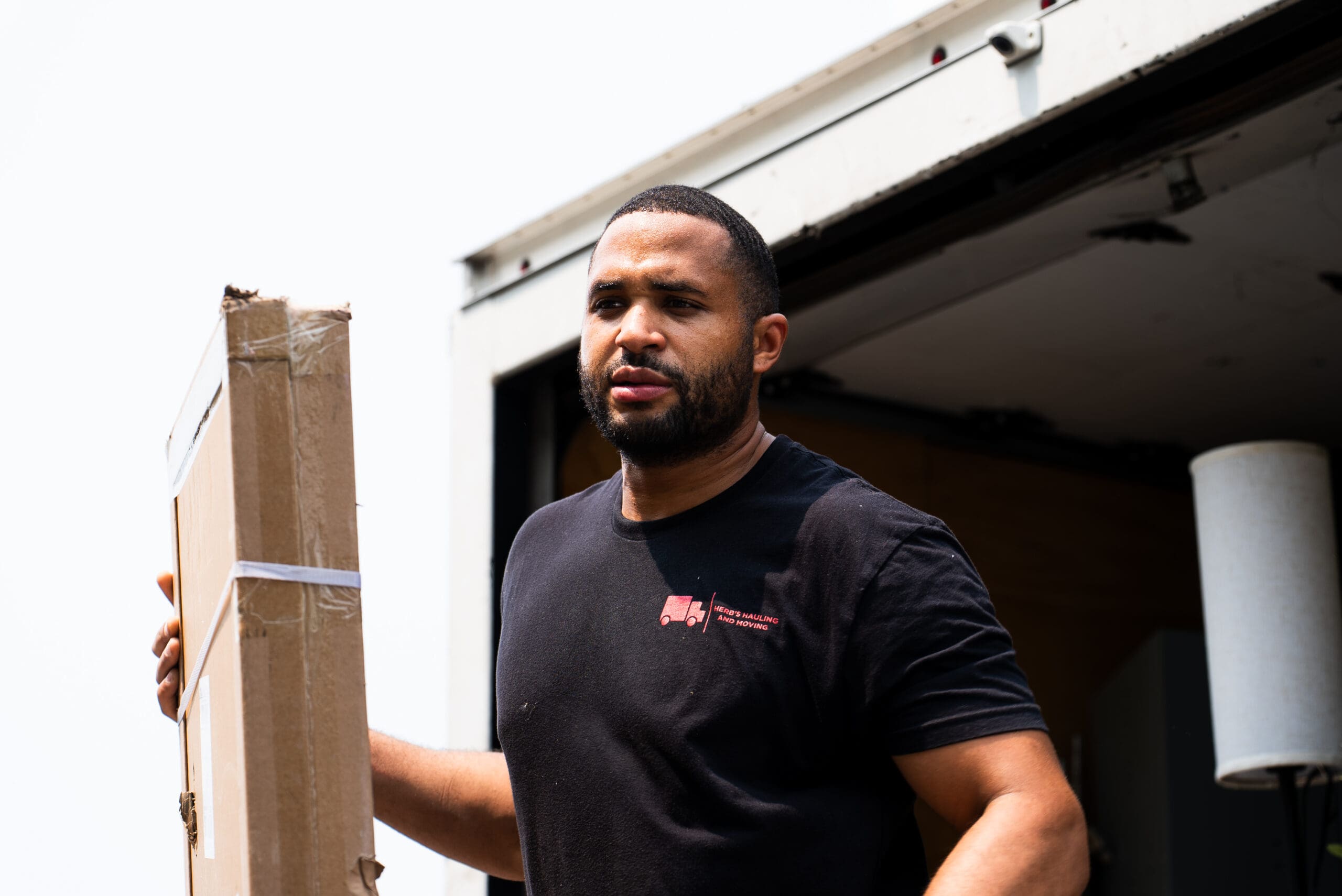 A man in a dark t-shirt demonstrates or instructs with a wooden beam in a garage or workshop setting.
